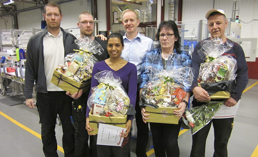 Sex personer står i en verkstadsmiljö, varav fyra håller i inslagna presentkorgar. De har uppmärksammats för Årets förbättringsförslag på AMB.
