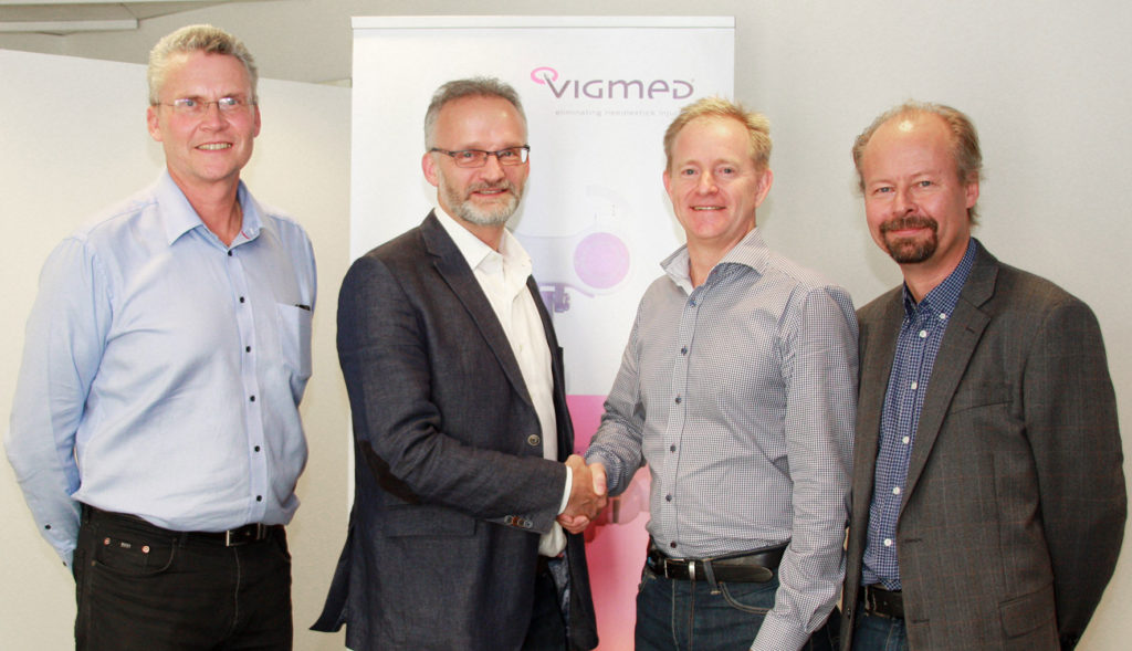 Four men in business attire posing in front of the Vigmed logo, with two of them shaking hands in front of the camera.
