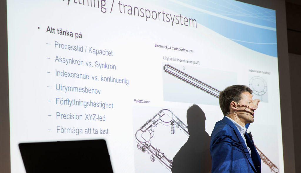 Föreläsare pekar på en presentation om transportsystem med punkter som processkapacitet, synkronisering och utrymmesbehov.