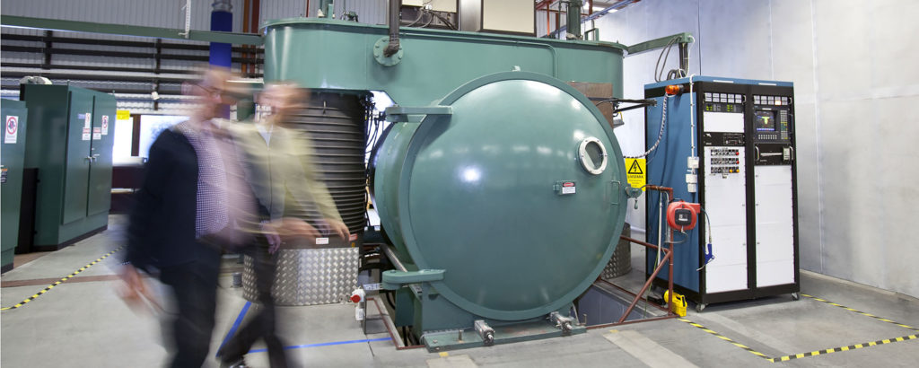 Två personer i rörelse framför en stor industriell vakuumugn i en produktionsmiljö med styrsystem och säkerhetsmarkeringar.