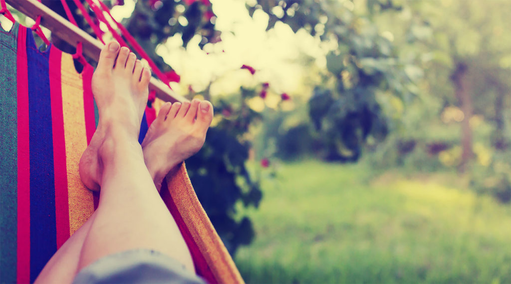 Person som ligger och slappnar av i en färgglad hängmatta utomhus med gröna träd i bakgrunden. Fokus på fötter i förgrunden.