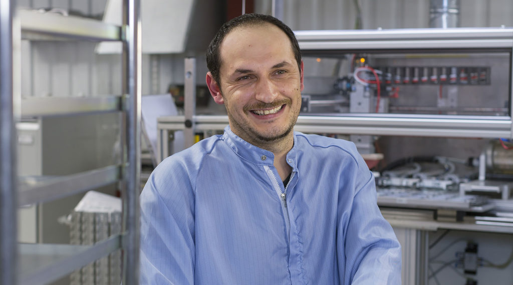 Leende man i blå antistatisk arbetsrock framför en maskin i en industriell produktionsmiljö.