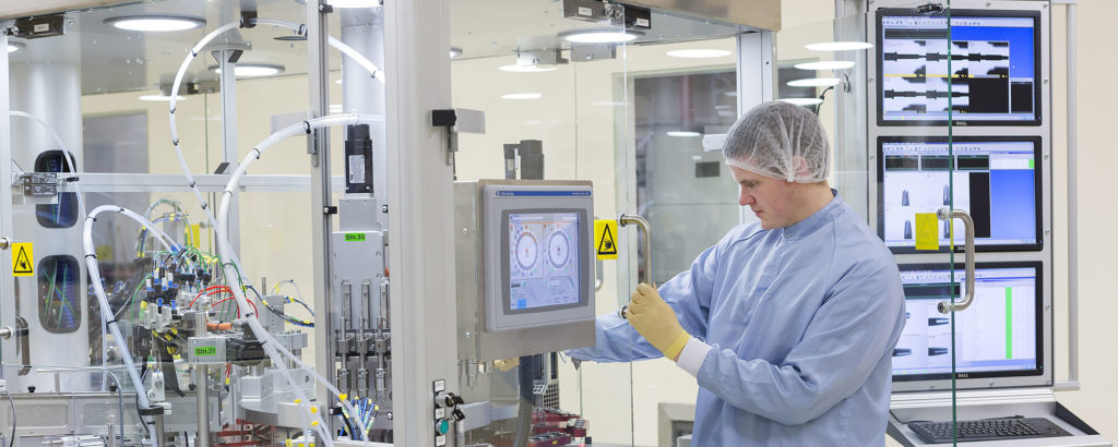 Processtekniker i renrumsmiljö som arbetar vid en avancerad maskin med pekskärm och automationsutrustning. Personen bär skyddskläder, mössa och handskar.