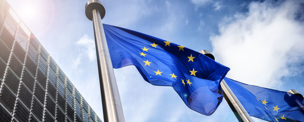 Europeiska unionens flagga som vajar i vinden framför en modern glasfasad och blå himmel med solreflexer.