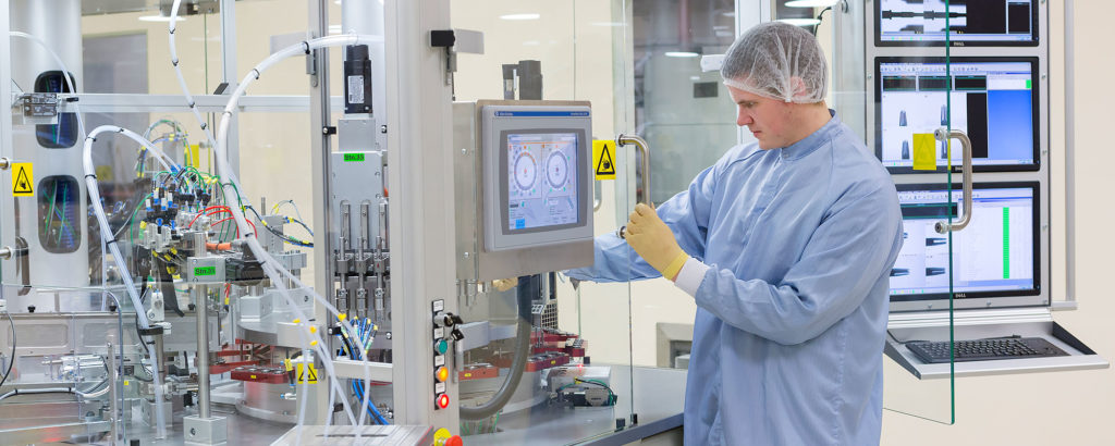 Processtekniker i renrumsmiljö som arbetar vid en avancerad maskin med pekskärm och automationsutrustning. Personen bär skyddskläder, mössa och handskar.