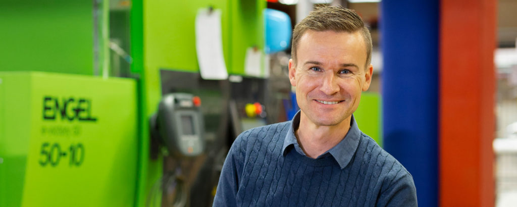 Peter Franzén, leende framför formsprutningsmaskiner i en färgstark produktionsmiljö.