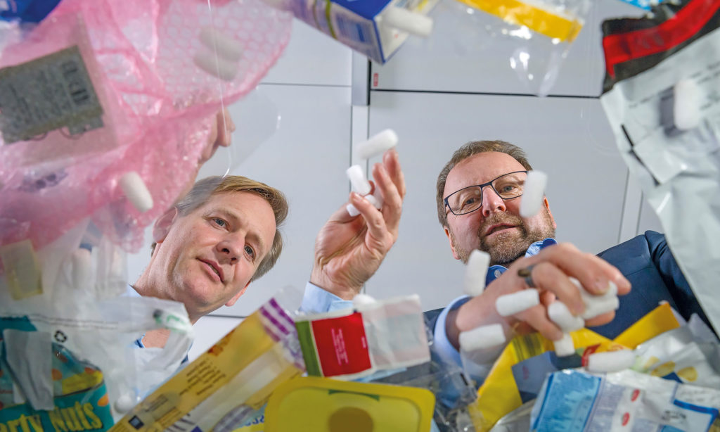 🇸🇪 Alt-text: Två män i kostym tittar ner i en hög med plastförpackningar och förpackningsmaterial, inklusive bubbelplast, plastpåsar och frigolit, i en miljö som liknar ett kontor eller laboratorium.