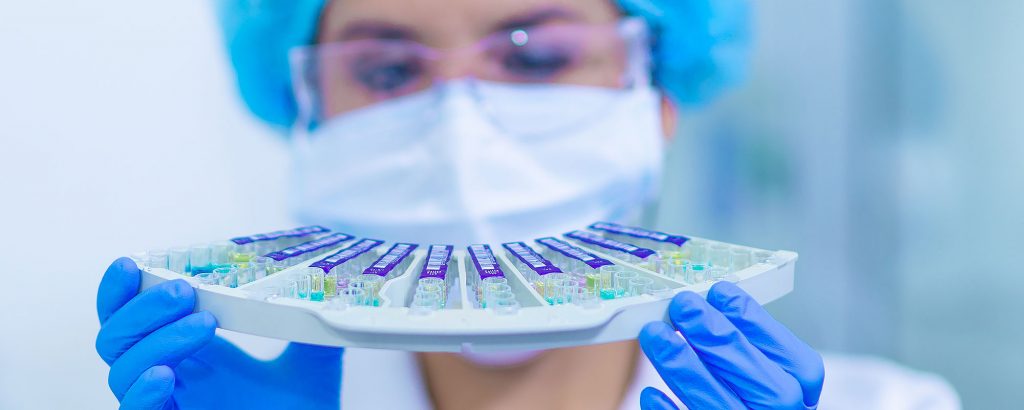 Närbild på en laboratoriearbetare med blå handskar som håller upp en bricka med färgade provrör – fokus på medicinteknik och diagnostik.
