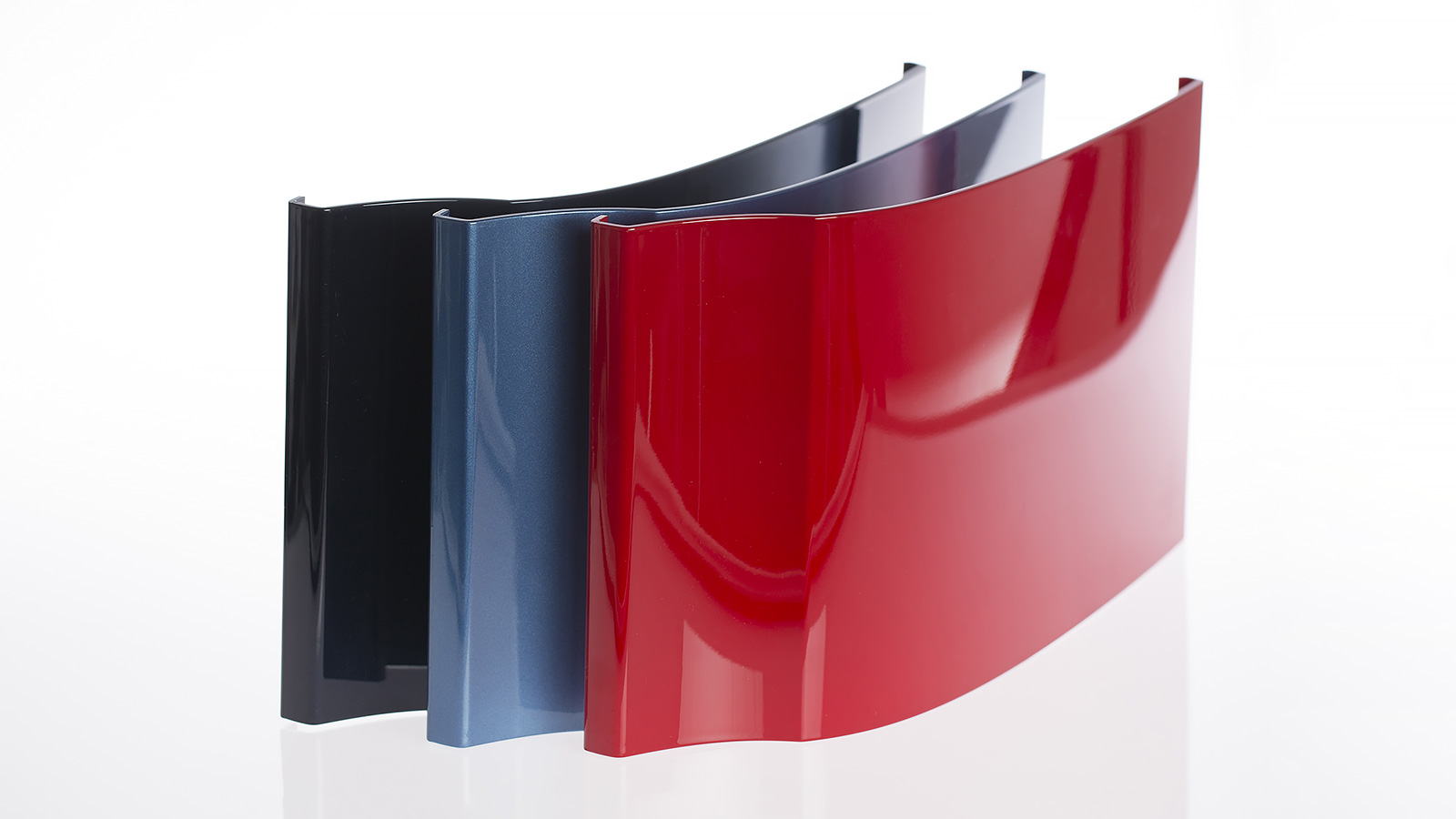 Three glossy, curved panels in black, blue, and red, arranged in a row on a white background – illustrating color and surface treatment tests.