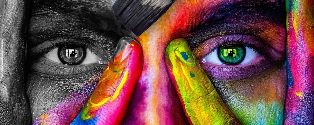 Närbild på ett ansikte där halva är i svartvitt och halva är målad i starka färger. En pensel sveper över pannan. Båda ögonen har speglingar av en person i ett fönster.