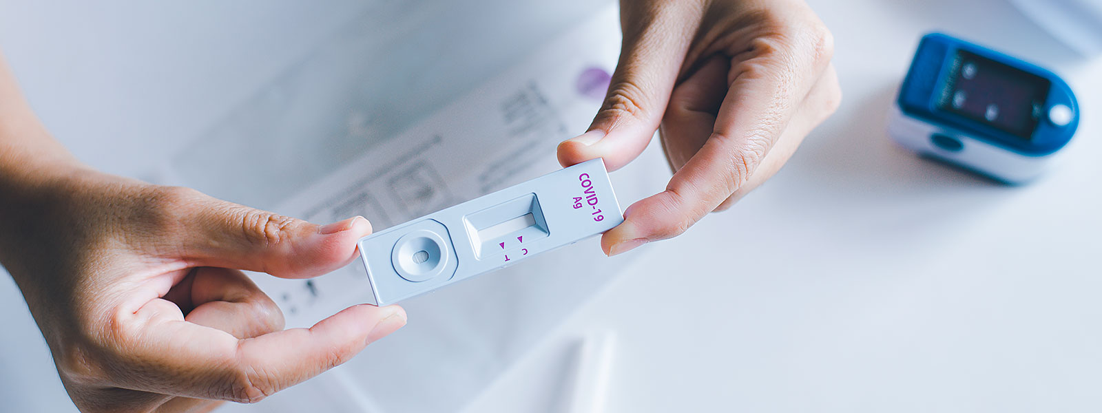 Närbild på händer som håller i ett COVID-19 antigentest för självtestning, med en pulsoximeter i bakgrunden.