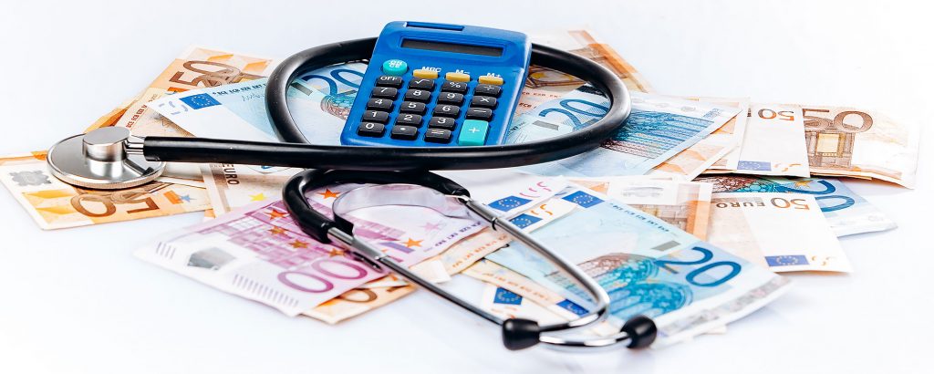 Stetoskop och miniräknare placerade ovanpå en hög med eurosedlar – symboliserar kostnader och ekonomi inom hälso- och sjukvård.