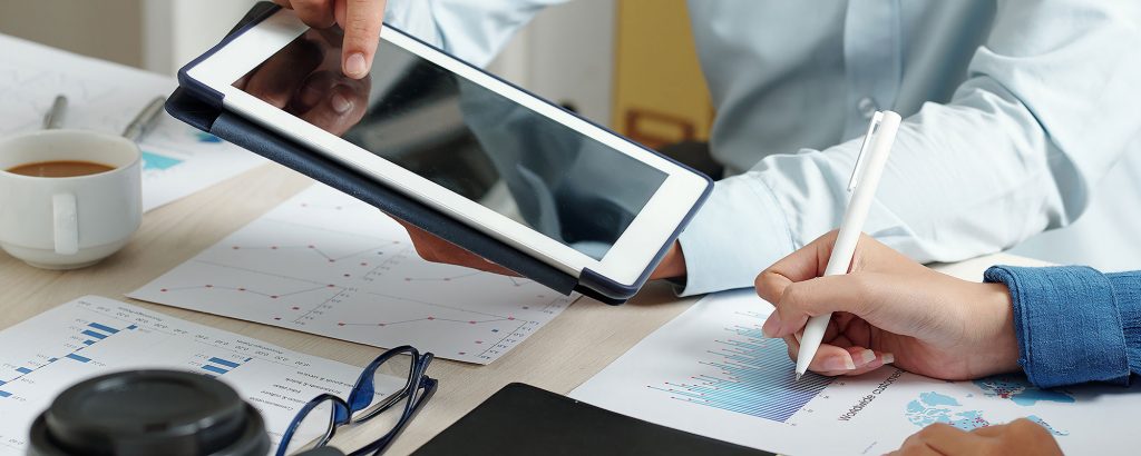 Två personer arbetar vid ett skrivbord med surfplatta, grafer och diagram, och antecknar i ett affärsdokument – fokus på analys och konceptutveckling.