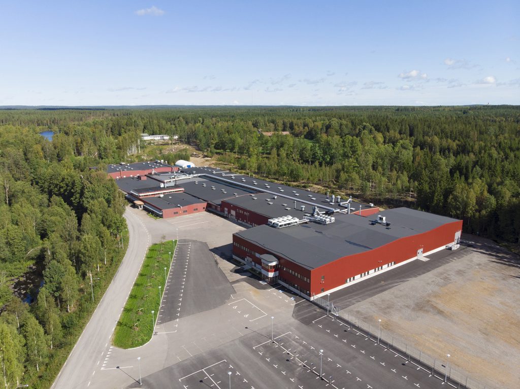 Fågelperspektiv över AMS stora produktionsanläggning med röda byggnader och parkeringsplatser, omgiven av skog.