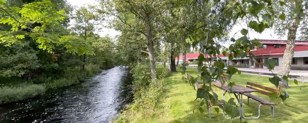 Lyckebyån som rinner genom ett grönområde intill AMB:s anläggning, med gräsmatta, träd och röda byggnader i bakgrunden.